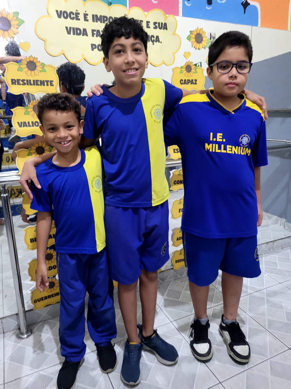 Três alunos do Instituto Educacional Millenium, de uniforme azul e amarelo, no corredor da escola